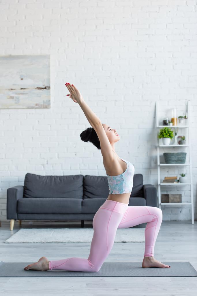vista lateral de la joven morena practicando media luna se abalanzan sobre la rodilla posan sobre esterilla de yoga - Foto, Imagen