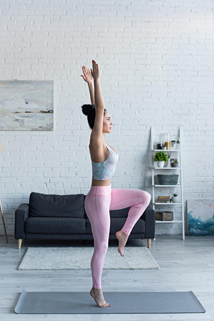 boční pohled na brunetku žena stojící na jedné noze se zvednutýma rukama při cvičení jógy - Fotografie, Obrázek