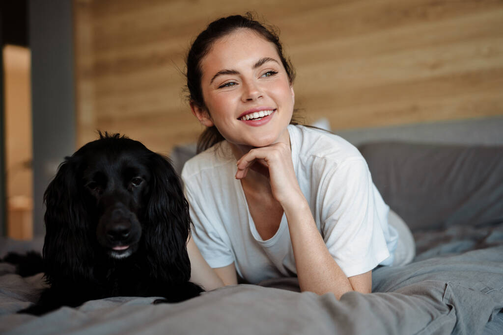 Χαμογελαστή μελαχρινή γυναίκα χαϊδεύει το σκύλο της στο σπίτι, ξαπλωμένη στον καναπέ - Φωτογραφία, εικόνα