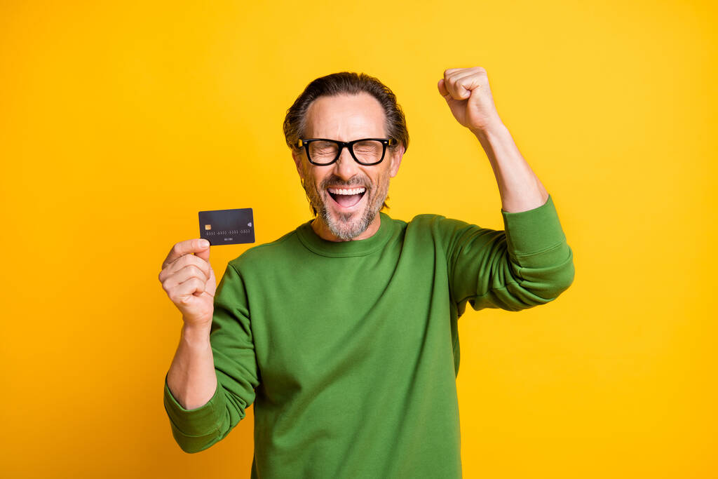 Kuva innoissaan mies esittää muovi pankkikortti nostaa nyrkki kulumista lasit vihreä villapaita eristetty keltainen väri tausta - Valokuva, kuva