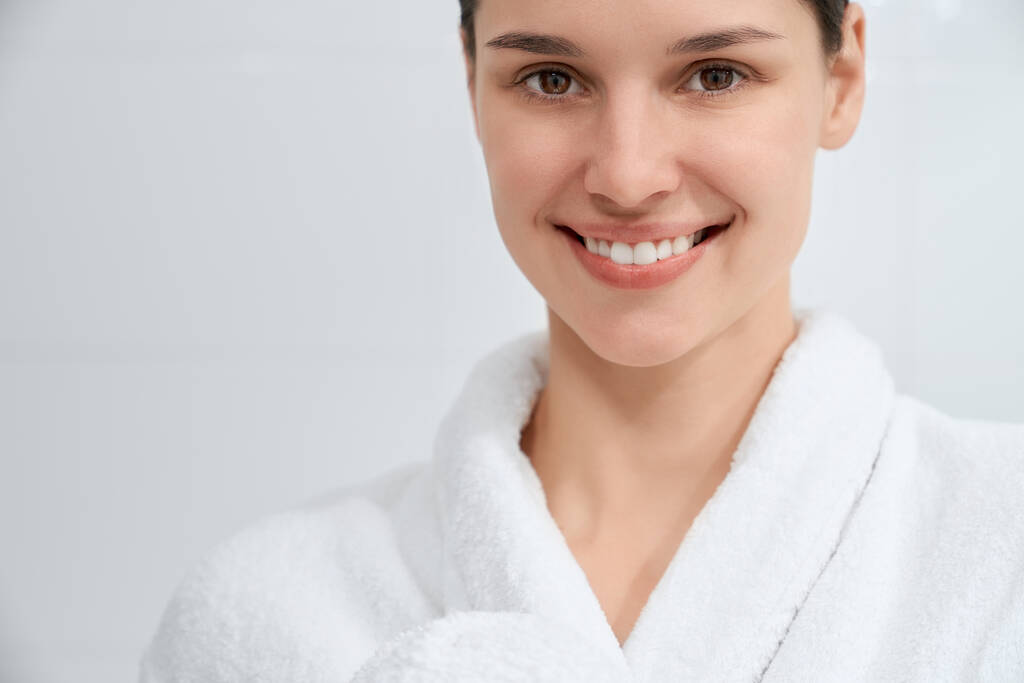 Sorridente donna attraente in vestaglia bianca in piedi in bagno. - Foto, immagini