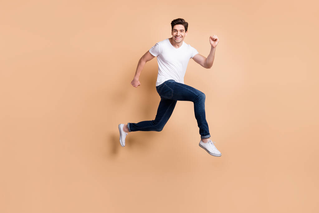 Full size profil zdjęcie hura brunet człowiek skok bieg nosić t-shirt jeansy trampki izolowane na beżowym tle - Zdjęcie, obraz