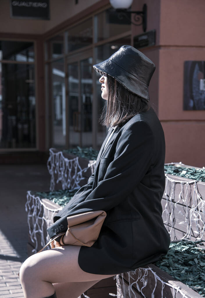 Una mujer con un sombrero negro, una chaqueta de hombre negro, y una falda muy corta se sienta en un banco en un día soleado de primavera. - Foto, imagen