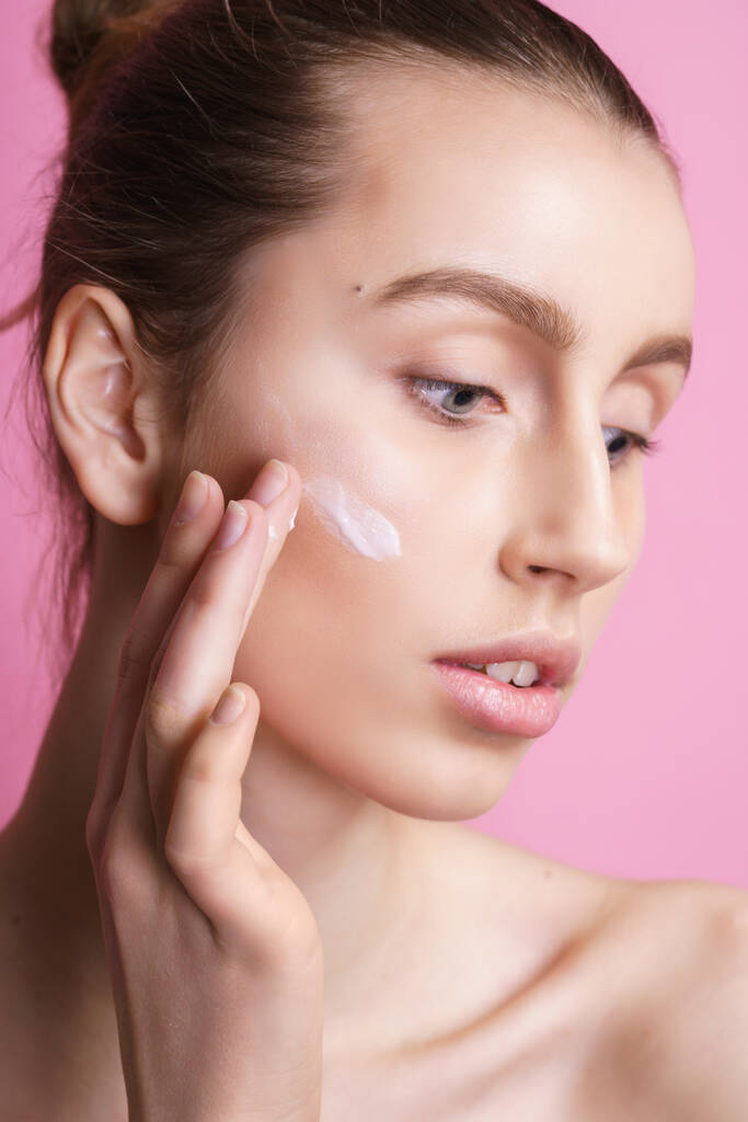 Mooie jonge vrouw aanbrengen crème op haar gezicht tegen kleur achtergrond - Foto, afbeelding