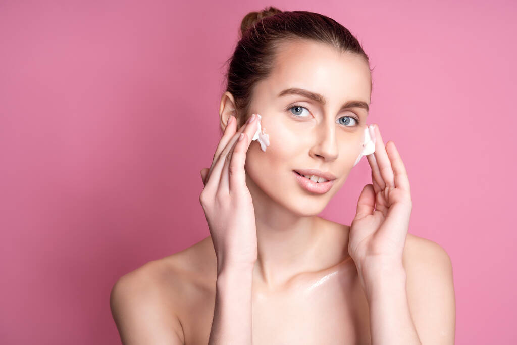 Mooie jonge vrouw aanbrengen crème op haar gezicht tegen kleur achtergrond - Foto, afbeelding
