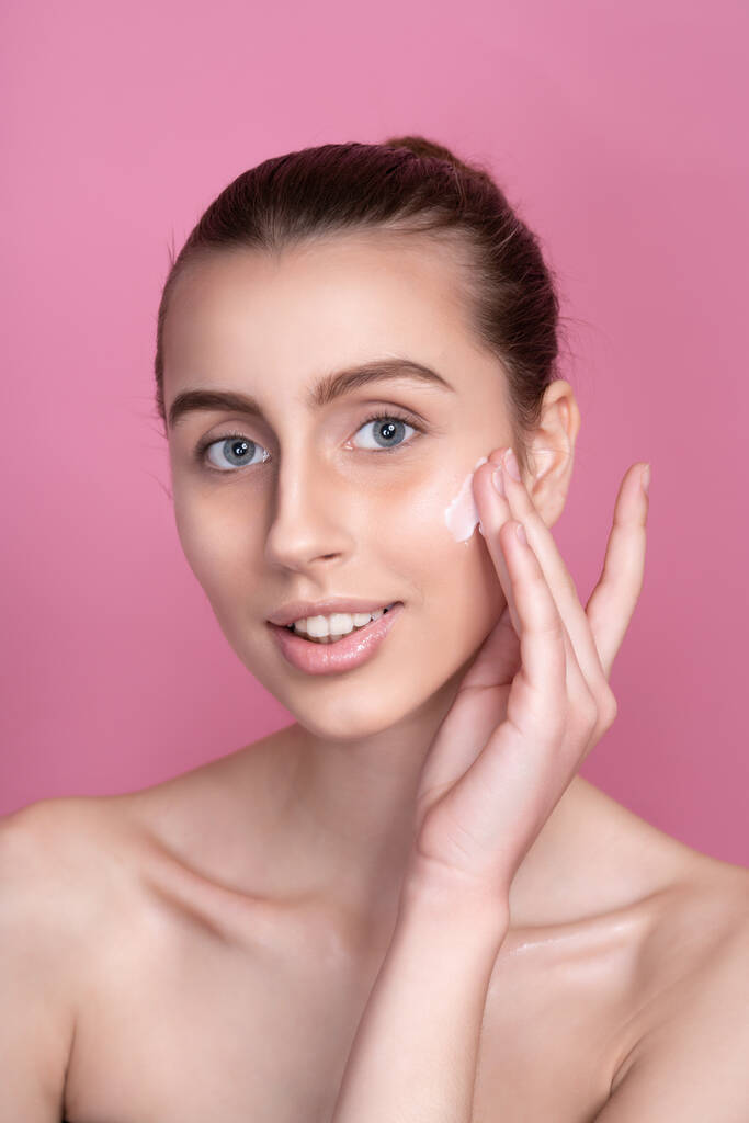 Hermosa mujer joven aplicando crema en su cara contra el fondo de color - Foto, Imagen