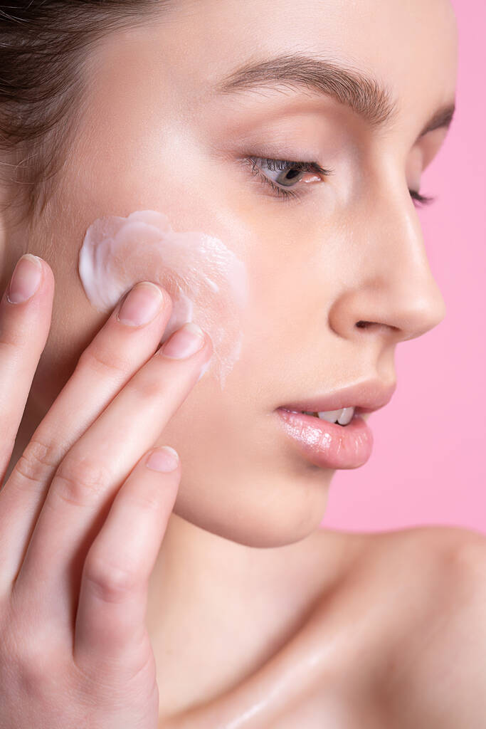 Hermosa mujer joven aplicando crema en su cara contra el fondo de color - Foto, Imagen
