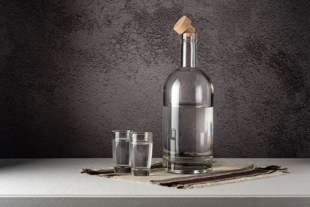 A glass bottle with glasses, a strong drink, on a white countertop against a gray wall - Foto, Imagen