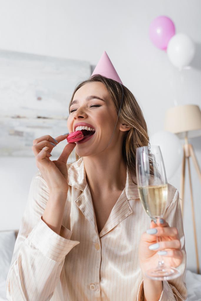Весела жінка в вечірній кепці їсть макаруни і тримає келих шампанського
  - Фото, зображення
