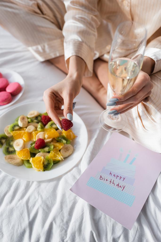 Vista cortada do cartão de aniversário perto da mulher com champanhe e salada de frutas na cama  - Foto, Imagem