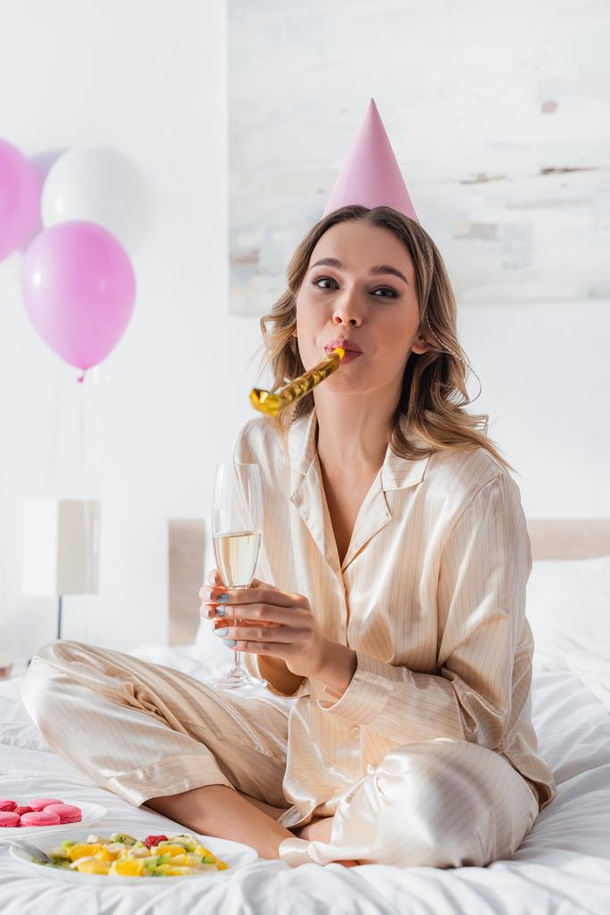 Mujer soplando cuerno de fiesta y la celebración de champán cerca de la comida en la cama durante la celebración del cumpleaños  - Foto, imagen