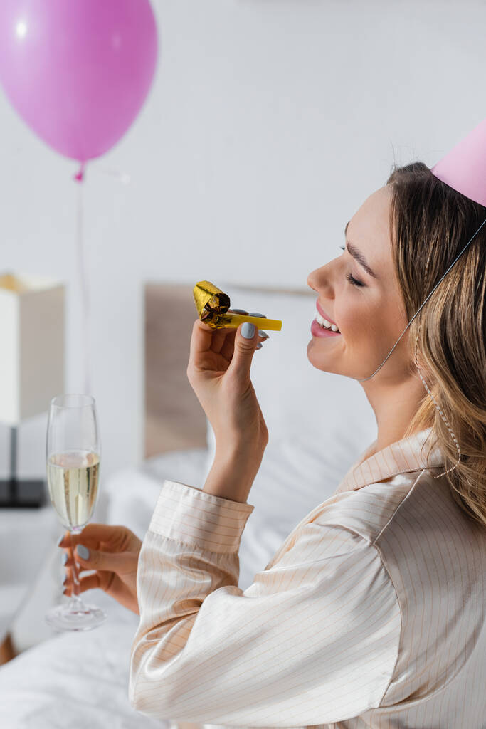 Zijaanzicht van de vrouw in party cap holding hoorn en champagne in de slaapkamer  - Foto, afbeelding