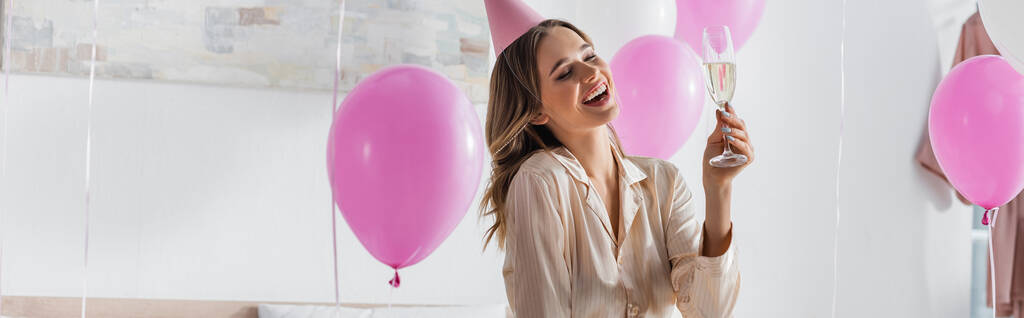Ευτυχισμένη γυναίκα με σαμπάνια γιορτάζει γενέθλια κοντά σε μπαλόνια στο σπίτι, πανό  - Φωτογραφία, εικόνα