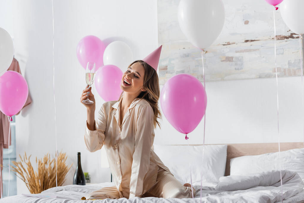 Счастливая женщина в шапочке для вечеринок держит бокал шампанского возле воздушных шаров в спальне  - Фото, изображение