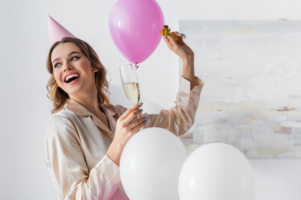 Glimlachende vrouw met feesthoorn en champagne vieren verjaardag in de buurt van ballonnen thuis  - Foto, afbeelding