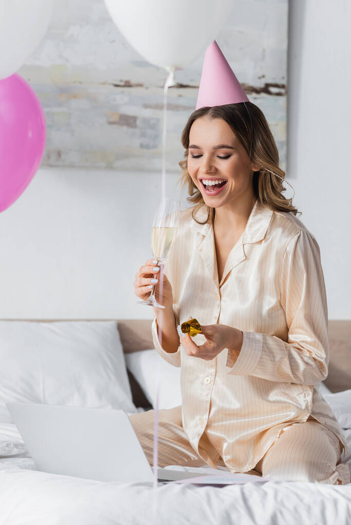Usmívající se žena v pyžamu drží šampaňské a party rohu u notebooku na posteli  - Fotografie, Obrázek