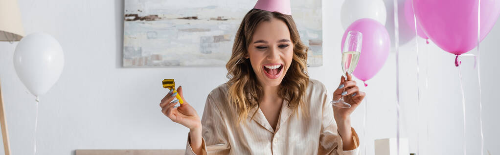 Счастливая женщина с рогом для вечеринок и шампанским празднует день рождения возле воздушных шаров дома, баннер  - Фото, изображение