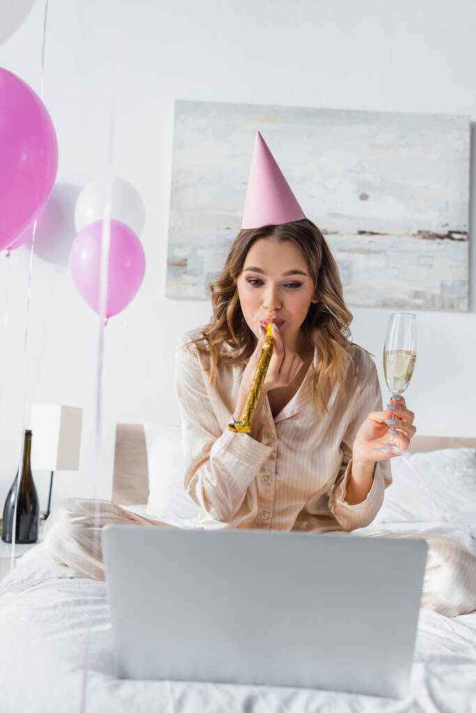 Woman with champagne blowing party horn near blurred laptop on bed  - Photo, Image