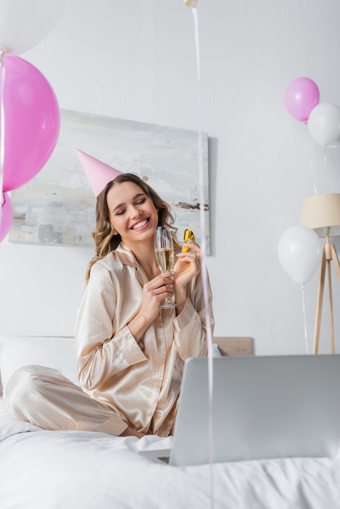 Mujer feliz con ventilador de fiesta, champán y gorra teniendo videollamada en el ordenador portátil durante el cumpleaños  - Foto, Imagen