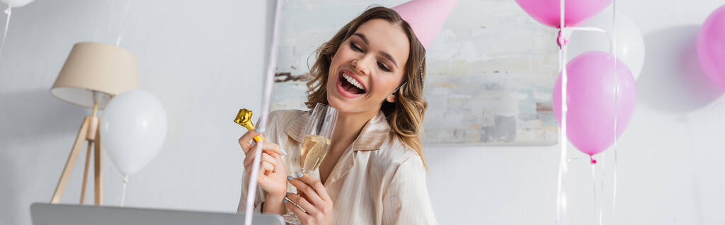 Mulher positiva com ventilador de festa e champanhe tendo chat de vídeo no laptop em casa, banner  - Foto, Imagem