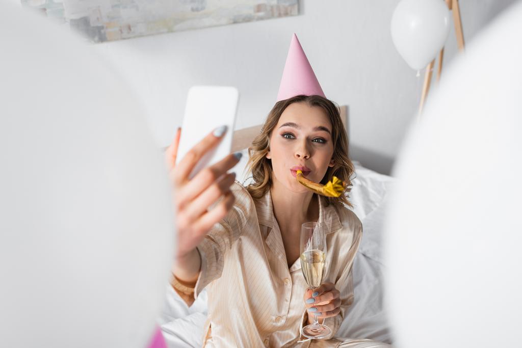 Жінка з партійним рогом і шампанським бере селфі під час вечірки на день народження в спальні
  - Фото, зображення