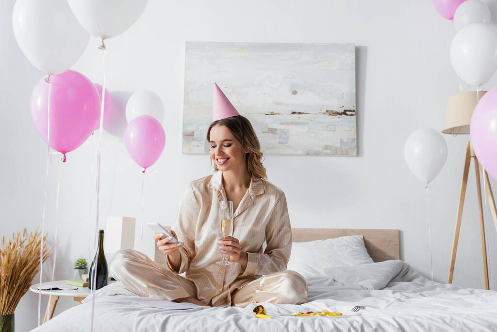 Glimlachende vrouw met champagne met smartphone in de buurt van fruitsalade en ballonnen in de slaapkamer  - Foto, afbeelding