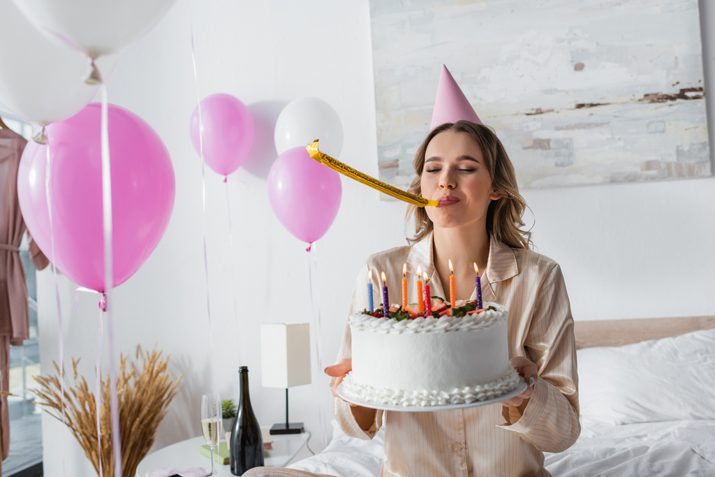 女性とともにパーティーホーンとキャップ保持誕生日ケーキ近く風船でベッドルーム  - 写真・画像