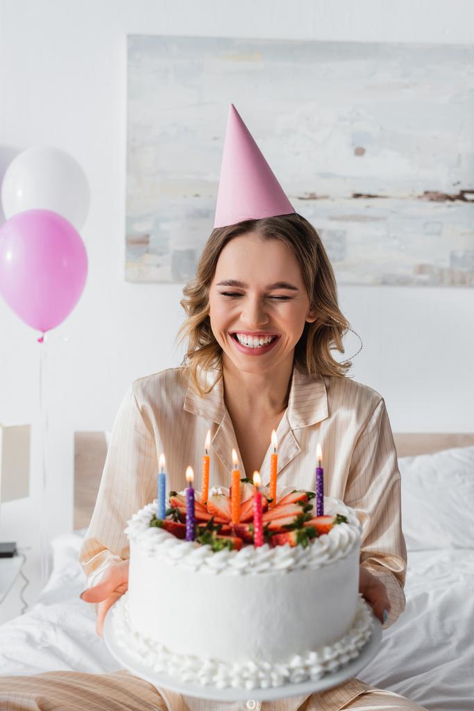 Positive woman in party cap holding birthday cake with candles in bedroom  - Photo, Image