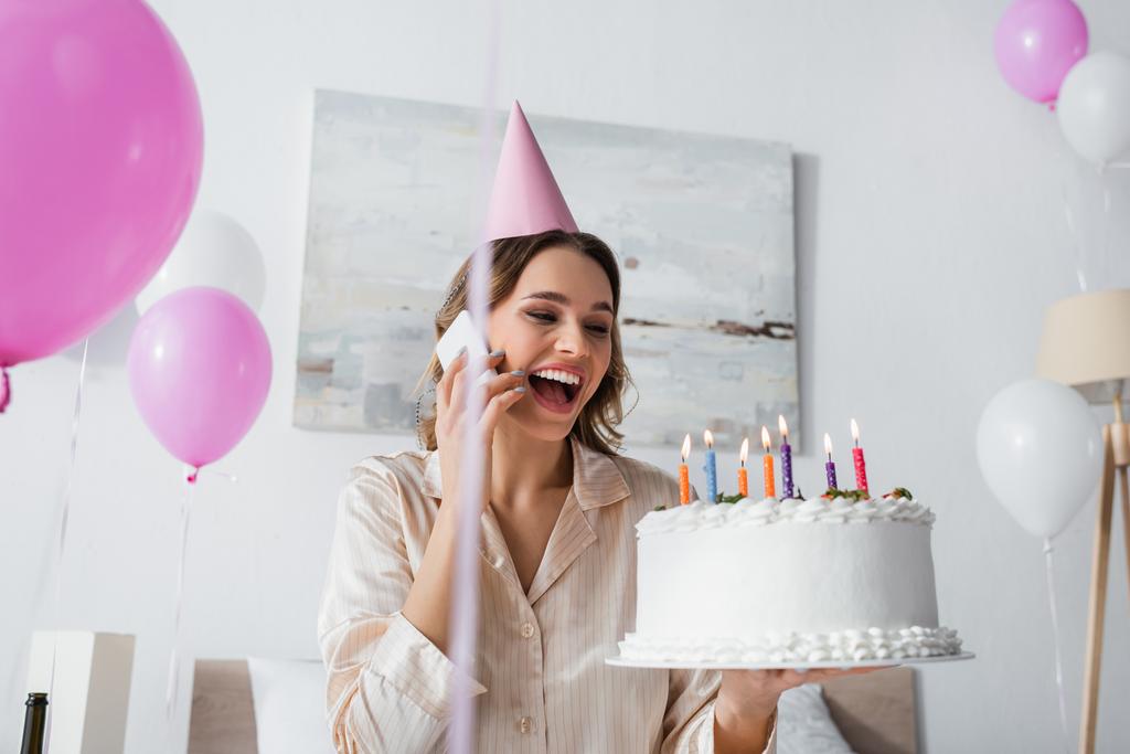 Femme avec gâteau d'anniversaire parlant sur smartphone et tenant gâteau d'anniversaire près de ballons à la maison  - Photo, image