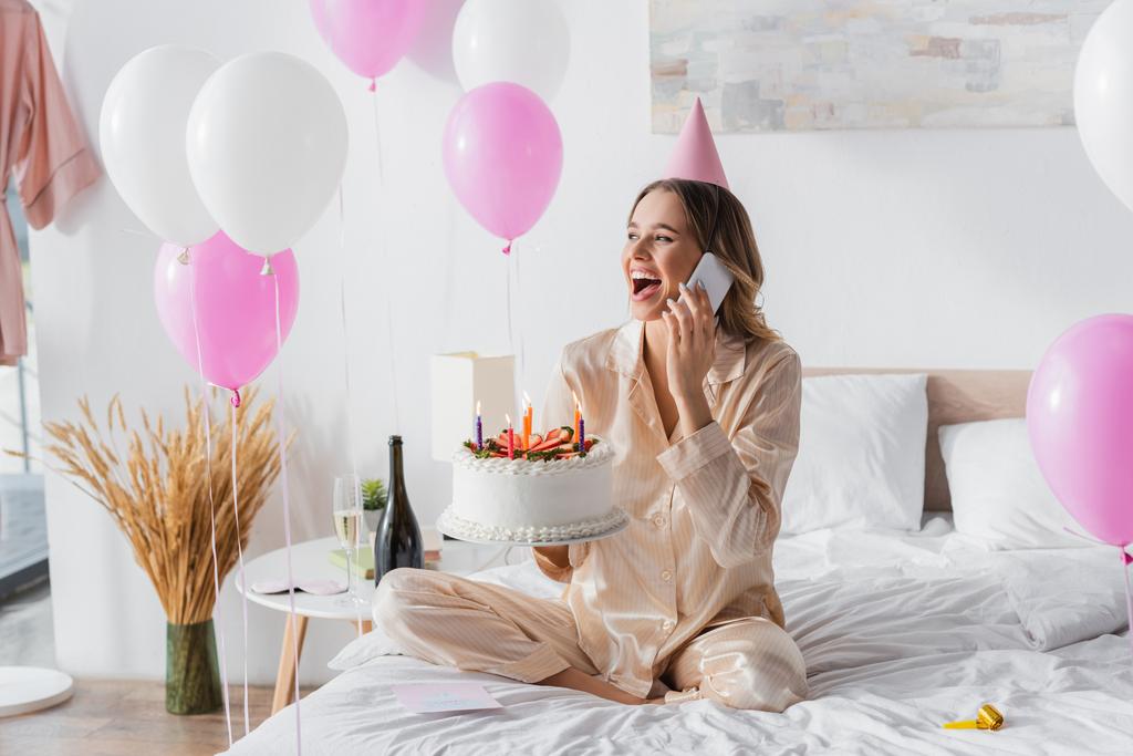 Mujer emocionada hablando por teléfono celular y sosteniendo pastel de cumpleaños con velas en la cama  - Foto, imagen