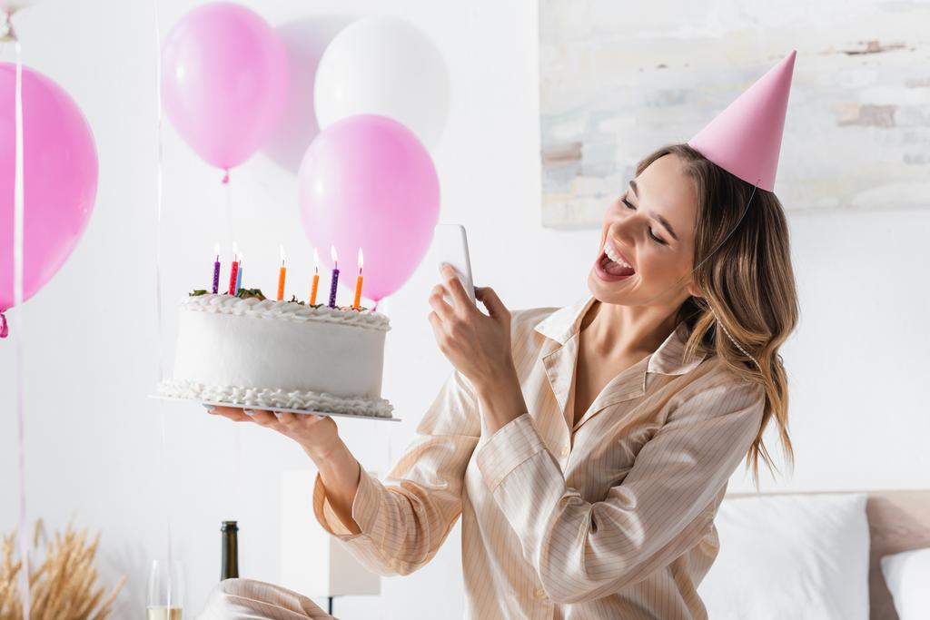 Cheerful woman with birthday cake taking photo on smartphone during party at home  - Photo, Image