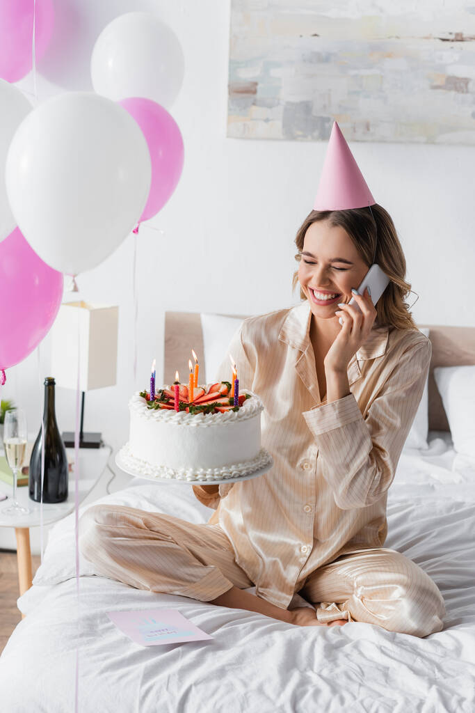 Mujer alegre hablando en el teléfono móvil y celebración de pastel de cumpleaños durante la celebración en el dormitorio  - Foto, imagen