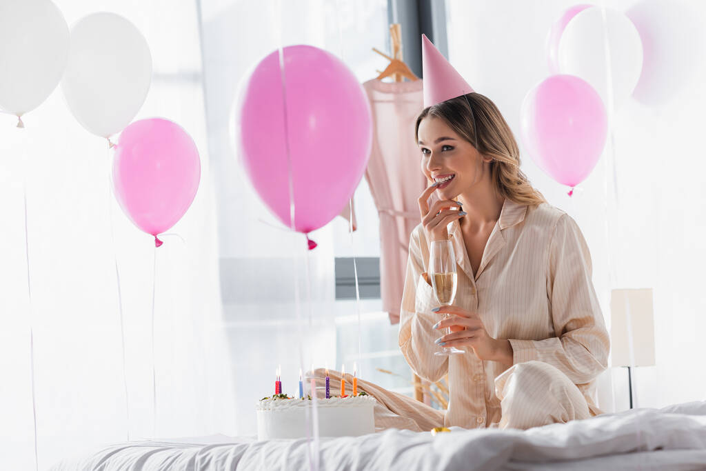 Lächelnde Frau mit Partymütze hält Champagner in der Nähe von Geburtstagstorte und Luftballons  - Foto, Bild