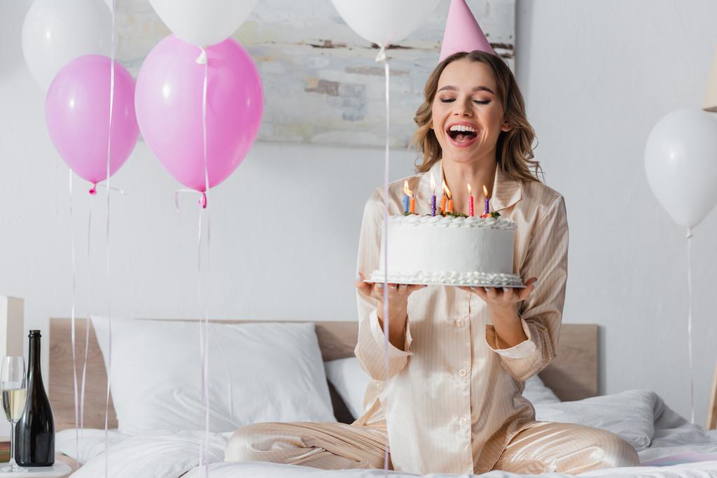 Ευτυχισμένη γυναίκα με καπέλο πάρτι κρατώντας τούρτα γενεθλίων κοντά σε μπαλόνια και σαμπάνια στην κρεβατοκάμαρα  - Φωτογραφία, εικόνα