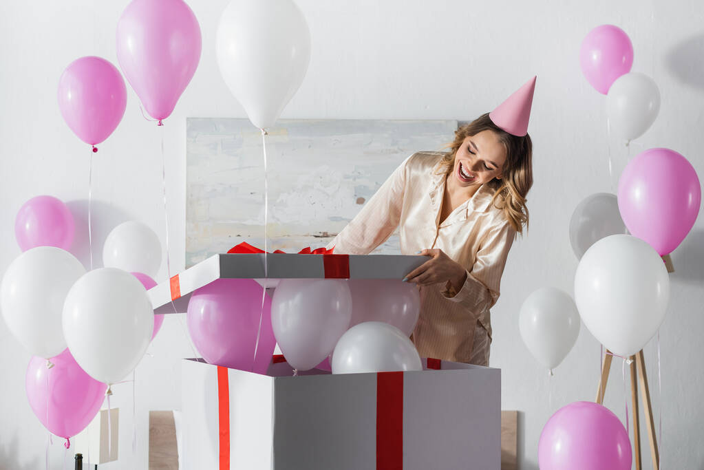 Femme en bonnet de fête et boîte cadeau d'ouverture de pyjama avec ballons festifs à la maison  - Photo, image