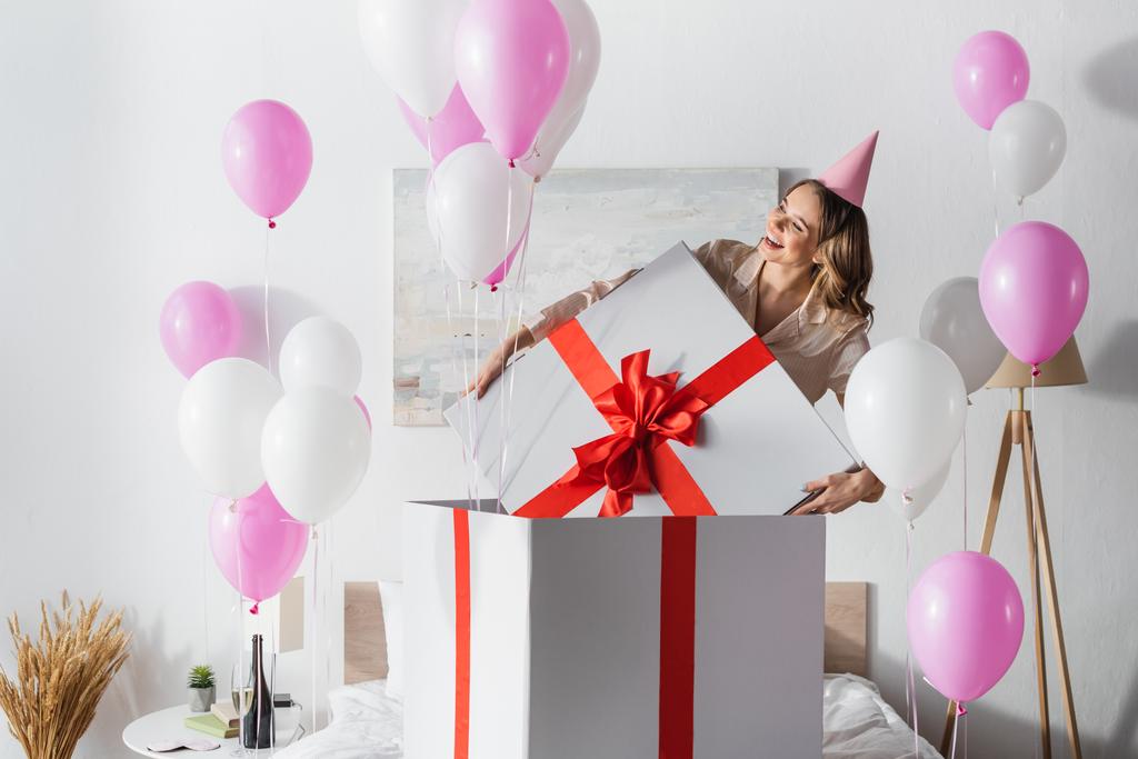 Mulher sorridente segurando boné de enorme presente perto de balões festivos no quarto  - Foto, Imagem