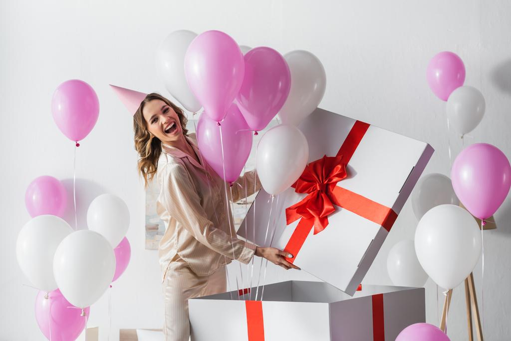 Позитивная женщина стоит рядом с большой подарочной коробкой с воздушными шарами во время празднования дня рождения дома  - Фото, изображение