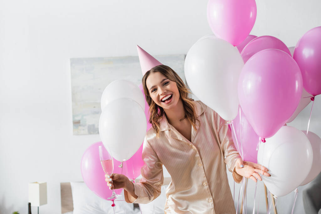 Pijamalı ve parti şapkalı neşeli kadın yatak odasındaki balonların yanında şampanya bardağı tutuyor.  - Fotoğraf, Görsel