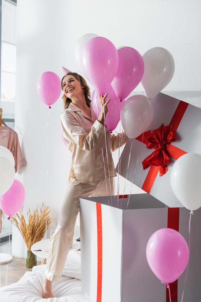 Mulher jovem abrindo enorme presente com balões festivos durante a festa de aniversário no quarto  - Foto, Imagem