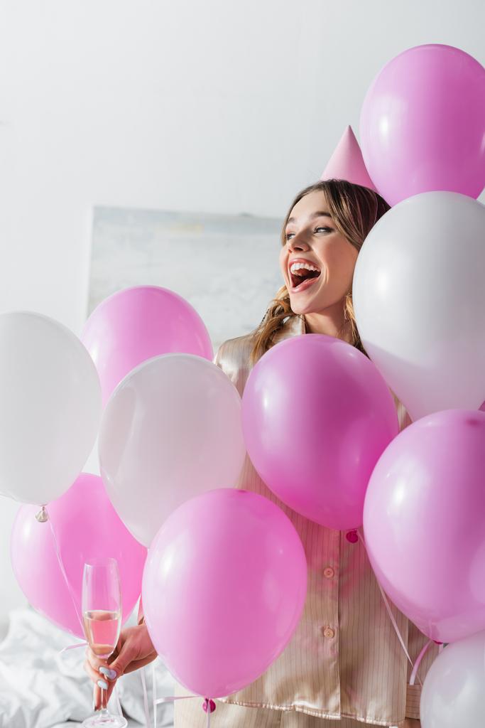 Aufgeregte Frau mit Champagner schaut bei festlichen Luftballons im Schlafzimmer weg  - Foto, Bild