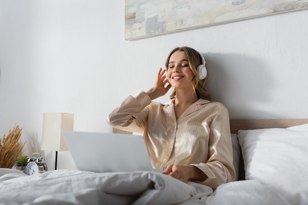 Cheerful woman listening music in headphones near blurred laptop on bed  - Photo, Image