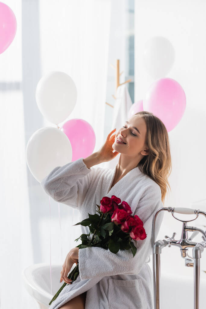 Positieve vrouw in badjas met rozen in de buurt van bad en ballonnen  - Foto, afbeelding