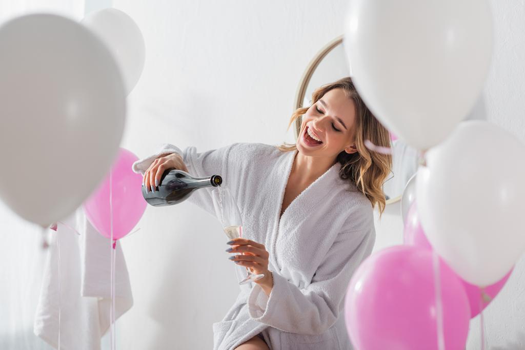Femme gaie en peignoir versant du champagne près des ballons dans la salle de bain  - Photo, image