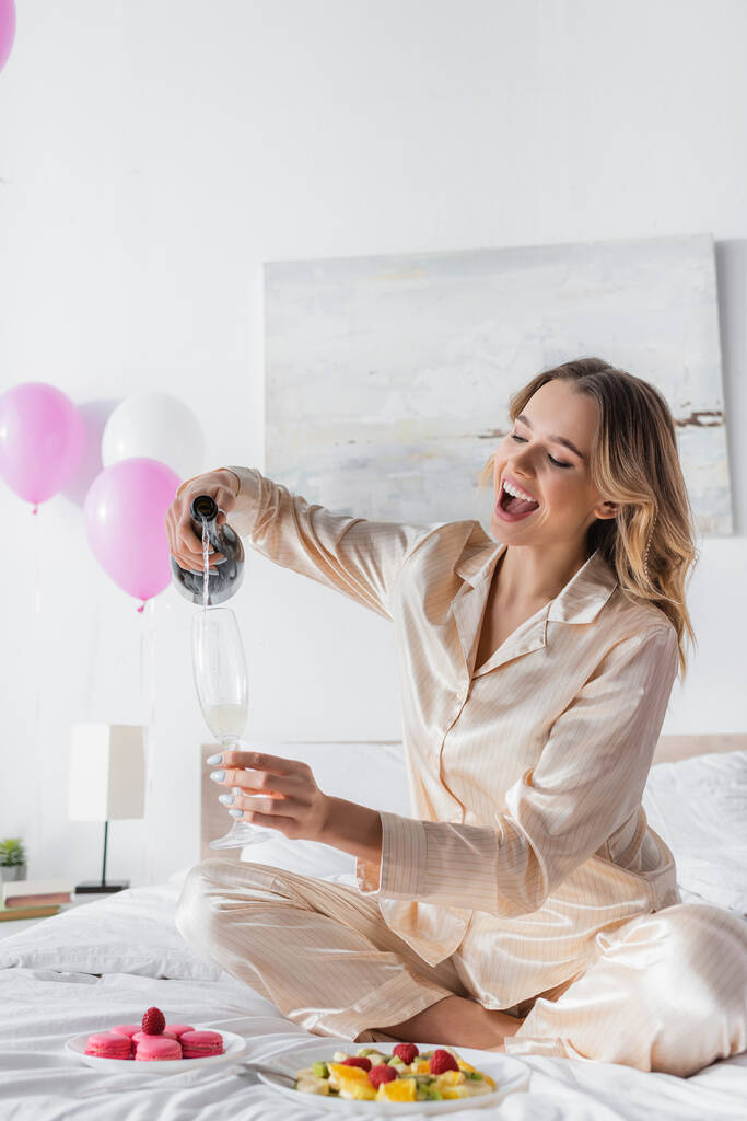 Woman pouring champagne near macaroons and fruit salad on bed  - Photo, Image