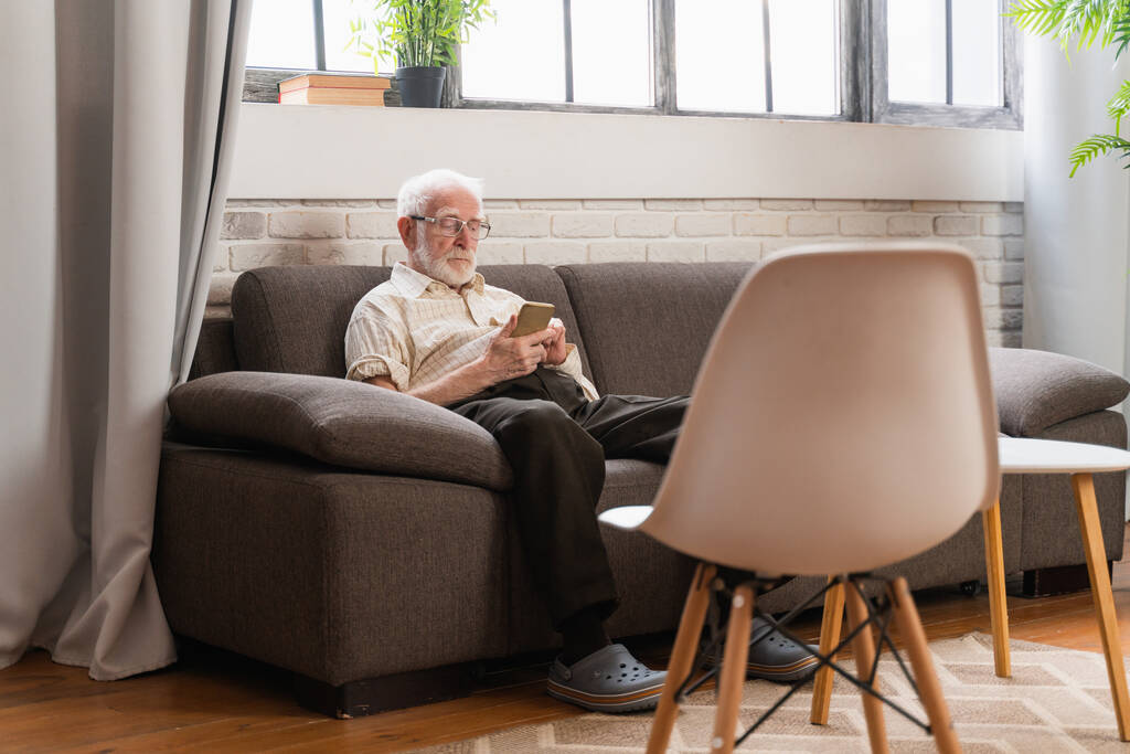 Старший мужчина сидит на диване смс на современном смартфоне - Старик один дома, видео-звонок своей семье с мобильного телефона - Фото, изображение