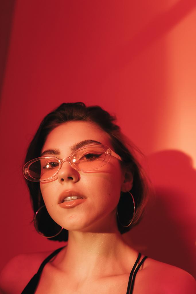 портрет молодої жінки в сонцезахисних окулярах дивиться на камеру на червоному тлі
 - Фото, зображення
