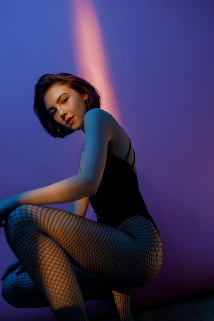young woman sitting in black bodysuit and fishnet tights in squat pose on violet and pink background - Photo, Image