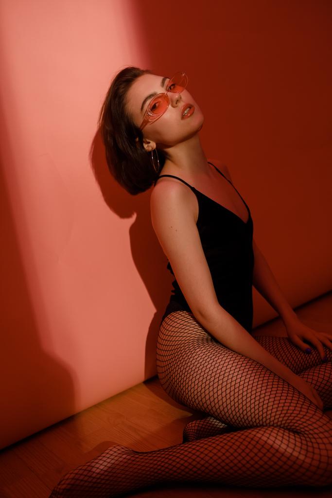 young woman sitting on floor in black bodysuit and fishnet tights on red background - Photo, Image