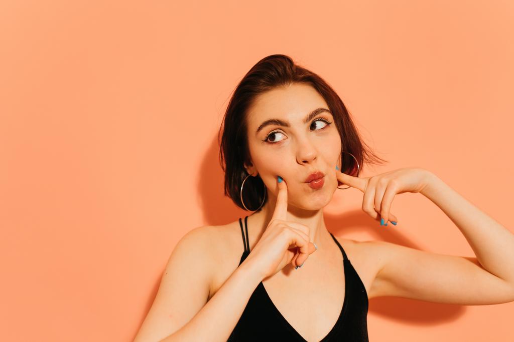 jonge vrouw flirten met pruilen lippen en handen in de buurt gezicht op oranje achtergrond - Foto, afbeelding