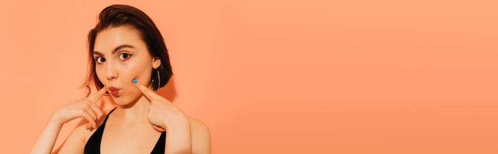 fiatal nő grimaszoló duzzadó ajkak és kezek közel arc narancs háttér, banner - Fotó, kép